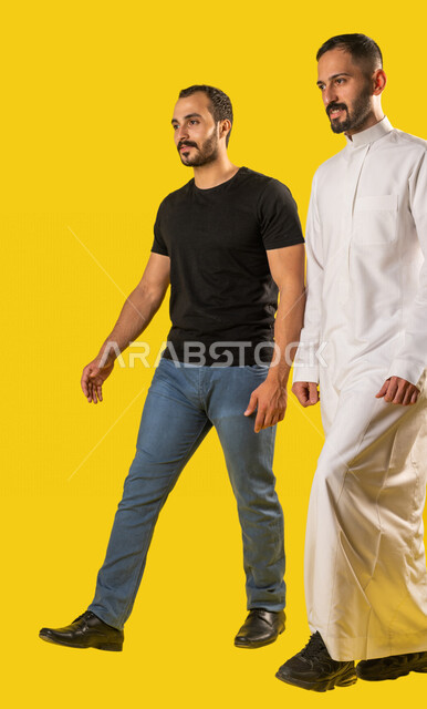 بورتريه لرجلان عربيان خليجيان سعوديان مبتسمان، صديقان يسيران معاً في نزهة ممتعة لإحدى الأماكن الترفيهية، صداقات وتعارف، علاقات إجتماعية، قضاء أوقات ممتعة، صورة الجسم بالطول الكامل، خلفية صفراء