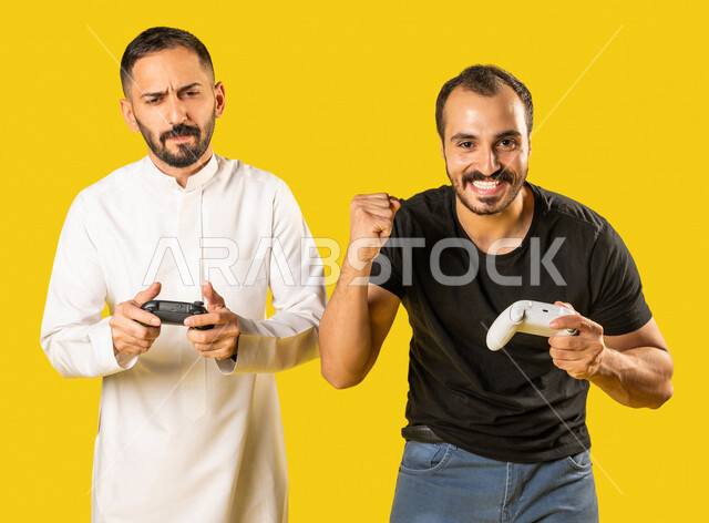 بورتريه لرجلان عربيان خليجيان سعوديان يحملان عصا التحكم، صديقان يستمتعان بلعب العاب الفيديو، اللعب والترفيه، التحدي والمغامرة، مفهوم الفوز والخسارة، تقنيات وألعاب، ايماءات وجه ويدين تدل على الفوز والانتصار، خلفية صفراء
