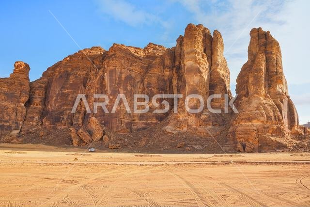 Rocky desert formations with sand in foreground, small car at distance, blue sky above, typical landscape of Al Ula, Saudi Arabia