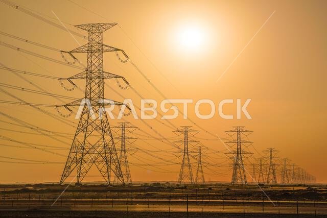 Large tall steel power pylons in rather flat landscape of Saudi Arabia, only black silhouettes visible against bright afternoon sun