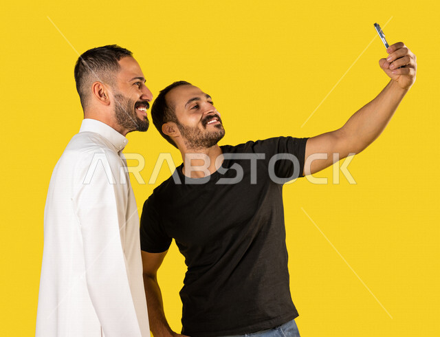 بورتريه لرجلان عربيان خليجيان سعوديان، التقاط صورة تذكارية من خلال الهاتف المحمول، صور تذكارية، صور سيلفي، كاميرا امامية، مكالمة فيديو، بث فيديو مباشر، ايماءات وجه ويدين تدل على السعادة، خلفية صفراء