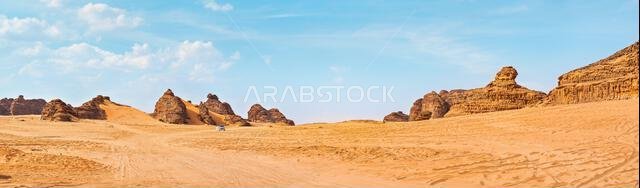 منظر صحراوي نموذجي في العلا، المملكة العربية السعودية، رمال تتخللها بعض الجبال، سيارة دفع رباعي صغيرة، رجل محلي وجمال على مسافة بعيدة