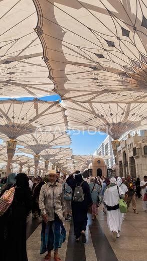 Medina, Saudi Arabia - March 6, 2024:  Pilgrims walk to the main mosque of the Prophet Muhammad Al-Masjid an-Nabawi underneath giant umbrella preparin