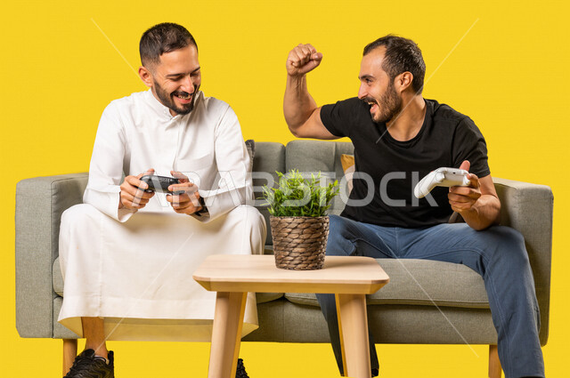 بورتريه لرجلان عربيان خليجيان سعوديان يحملان عصا التحكم، صديقان يستمتعان بلعب العاب الفيديو، اللعب والترفيه، التحدي والمغامرة، مفهوم الفوز والخسارة، تقنيات وألعاب، ايماءات وجه ويدين تدل على الفوز والانتصار، خلفية صفراء