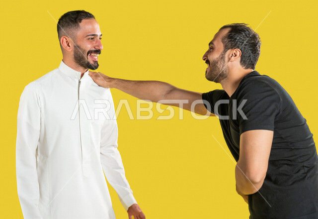 بورتريه لرجلان عربيان خليجيان سعوديان، العلاقات الأخوية المترابطة، علاقات اجتماعية، تعارف وصداقات، صديق يضع يده على كتف صديقه، إيماءات وجه ويدين تدل على السعادة الشديدة، خلفية صفراء