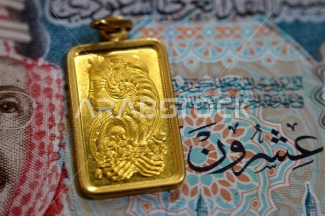 A closeup view of Saudi Arabian riyals money banknote and Lady Fortuna motif on a gold bar pendant, 24K Gold Bar, pure fine gold 999.9, for investment