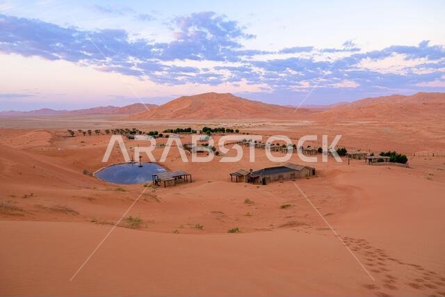 Remote desert camp featuring tents and buildings surrounding a refreshing oasis pond, nestled among vast sand dunes in the Empty Quarter of Oman