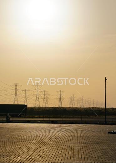 Large tall steel power pylons in rather flat landscape of Saudi Arabia, only black silhouettes visible against bright afternoon sun