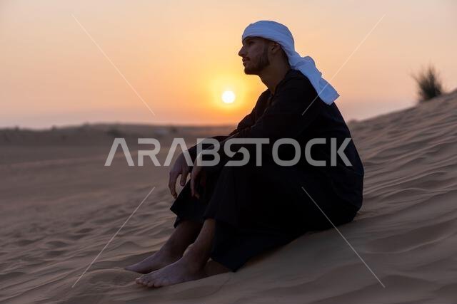 رجل عربي يجلس على الرمال في الصحراء وقت الغروب، مرتدياً الزي العربي التقليدي. صورة عالية الجودة