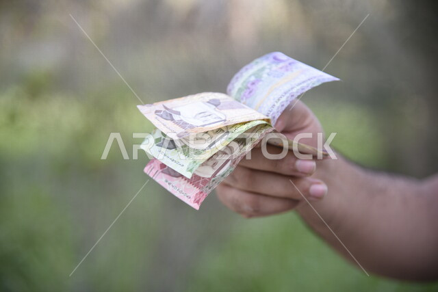 صورة مقربة ليد رجل يحمل اوراق نقدية من عملة الريال السعودي، عملة ورقية، مالية واقتصاد، العملة الرسمية للسعودية، الربح المالي، الاستثمار الاقتصادي، التنمية الاقتصادية، زكاه المال، زكاة المحاصيل الزراعية، خلفية للطبيعة