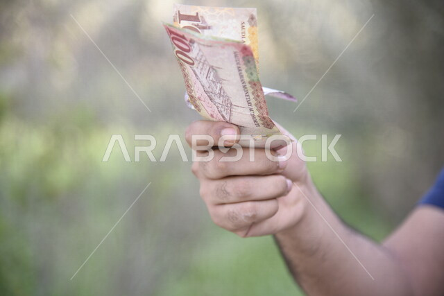صورة مقربة ليد رجل يحمل اوراق نقدية من عملة الريال السعودي، عملة ورقية، مالية واقتصاد، العملة الرسمية للسعودية، الربح المالي، الاستثمار الاقتصادي، التنمية الاقتصادية، زكاه المال، زكاة المحاصيل الزراعية، خلفية للطبيعة