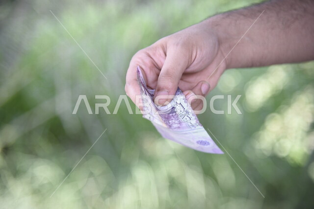 صورة مقربة ليد رجل يحمل اوراق نقدية من عملة الريال السعودي، عملة ورقية، مالية واقتصاد، العملة الرسمية للسعودية، الربح المالي، الاستثمار الاقتصادي، التنمية الاقتصادية، زكاه المال، زكاة المحاصيل الزراعية، خلفية للطبيعة
