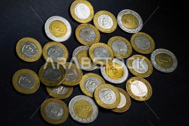 Saudi Arabian one and two riyal coins scattered on a dark surface, showing modern bimetallic design and Arabic inscriptions.