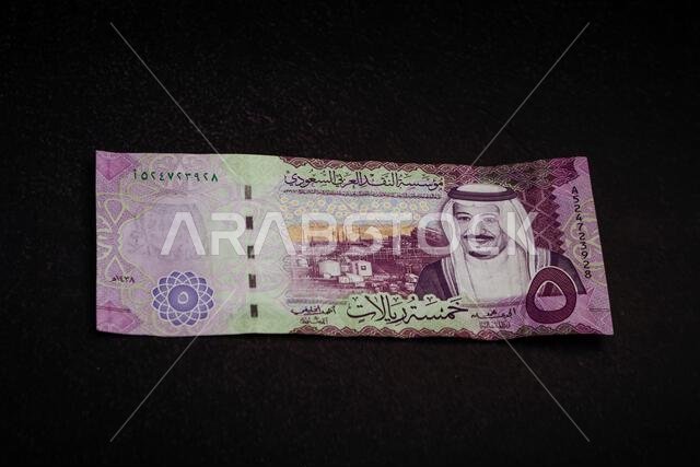 أوراق نقدية من فئة الريال السعودي على خلفية داكنة تحمل صور الملك سلمان ومعالم وطنية سعودية.
