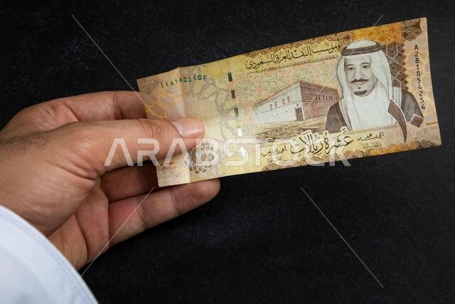 يد تحمل ورقة نقدية من الريال السعودي، تظهر صورة الملك سلمان وتصميم قصر الحكم في الرياض.