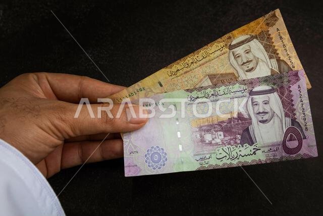 يد تحمل ورقة نقدية من الريال السعودي، تظهر صورة الملك سلمان وتصميم قصر الحكم في الرياض.