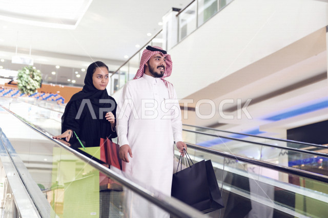 A Saudi Arabian Gulf couple on the escalator happy shopping together, the couple participating in shopping choices and exchanging opinions, enjoying buying and shopping, taking advantage of offers and discounts, spending a good time shopping from the mall