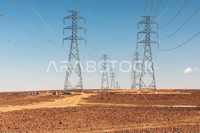 Two rows of towering electricity pylons traverse a barren desert, connecting distant power networks. The scene highlights infrastructure, scale, and