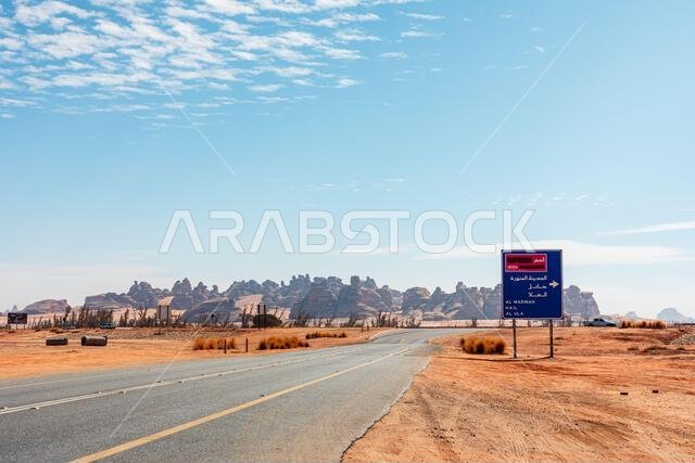 طريق صحراوي ناءٍ يؤدي إلى الحِجر في المملكة العربية السعودية، يشق طريقه عبر الرمال الحمراء، مع لافتة زرقاء وسط تكوينات صخرية وعرة. سماء صافية، طريق هادئ، ومساحة شاسعة