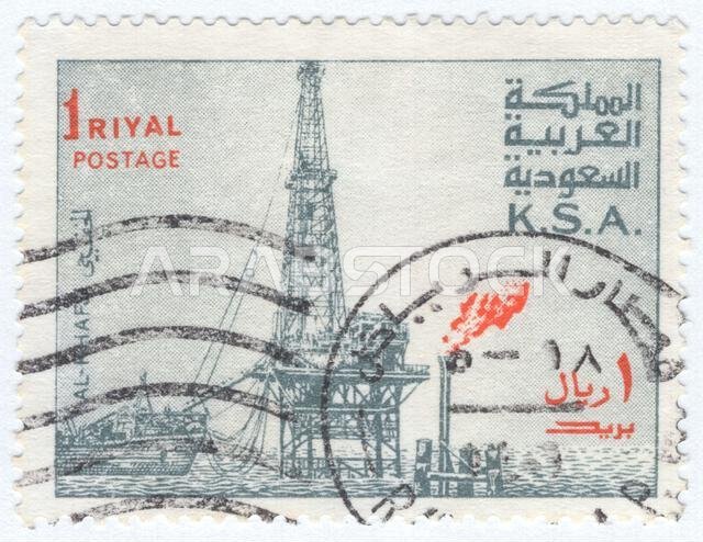 المملكة العربية السعودية - ١٩٧٧: طابع بريدي بقيمة ريال واحد، رمادي وبرتقالي مخضر، يصور مشهدًا صناعيًا لمنصة حفر نفطية بحرية في حقل الخفجي. يظهر برج حفر فولاذي ضخم يرتفع من منصة في البحر، مع سفينة خدمات راسية بجانبه إلى اليسار. ويظهر شعلة غاز مشتعلة باللون البرتقالي الفاقع على اليمين، في تناقض مع اللون الرمادي الباهت لبقية الآلات. المملكة العربية السعودية - ١٩٧٧: طابع بريدي بقيمة ريال واحد، رمادي وبرتقالي مخضر، يصور مشهدًا صناعيًا لمنصة حفر نفطية بحرية في حقل الخفجي. يظهر برج حفر فولاذي ضخم يرتفع من منصة في البحر، مع سفينة خدمات راسية بجانبه إلى اليسار. ويظهر شعلة غاز مشتعلة باللون البرتقالي الفاقع على اليمين، في تناقض مع اللون الرمادي الباهت لبقية الآلات.