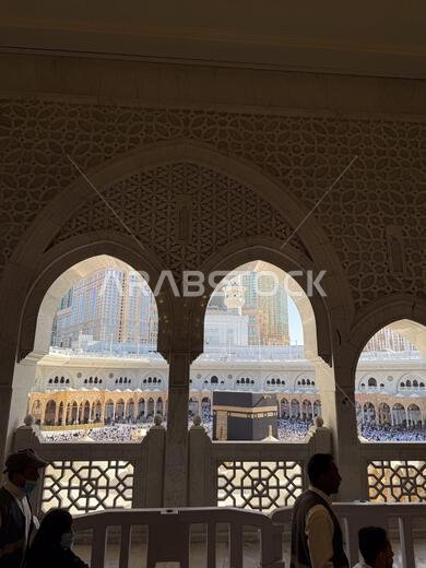منظر للكعبة والحجاج في المسجد الحرام بمكة المكرمة، محاط بأقواس المسجد المزخرفة.