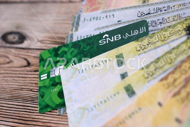 كييف، أوكرانيا - ٢٨ يناير ٢٠٢٦: صورة مقرّبة لبطاقة البنك الوطني السعودي وأوراق نقدية من الريال السعودي على طاولة. يُعدّ البنك الوطني السعودي البنك الأكثر شعبية في المملكة العربية السعودية.