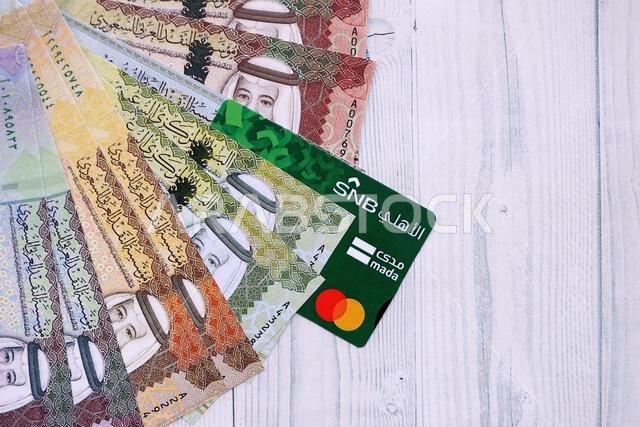 كييف، أوكرانيا - ٢٨ يناير ٢٠٢٦: صورة مقرّبة لبطاقة البنك الوطني السعودي وأوراق نقدية من الريال السعودي على طاولة. يُعدّ البنك الوطني السعودي البنك الأكثر شعبية في المملكة العربية السعودية.