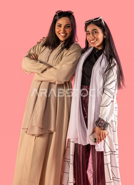 Portrait of a smiling Saudi Arabian Gulf mother with folded hands standing next to her happy young daughter to take a souvenir photo, a photo for remembrance, a mother and her daughter are friends, happy family family relationships, pink background