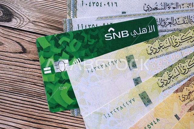 كييف، أوكرانيا - ٢٨ يناير ٢٠٢٦: صورة مقرّبة لبطاقة البنك الوطني السعودي وأوراق نقدية من الريال السعودي على طاولة. يُعدّ البنك الوطني السعودي البنك الأكثر شعبية في المملكة العربية السعودية.