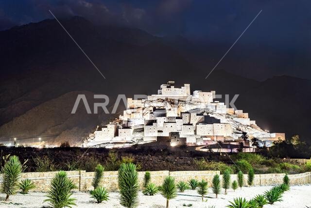 The illuminated stone houses of Thee Ain Heritage Village at night against the backdrop of the Sarawat Mountains in Al Bahah, Saudi Arabia.
