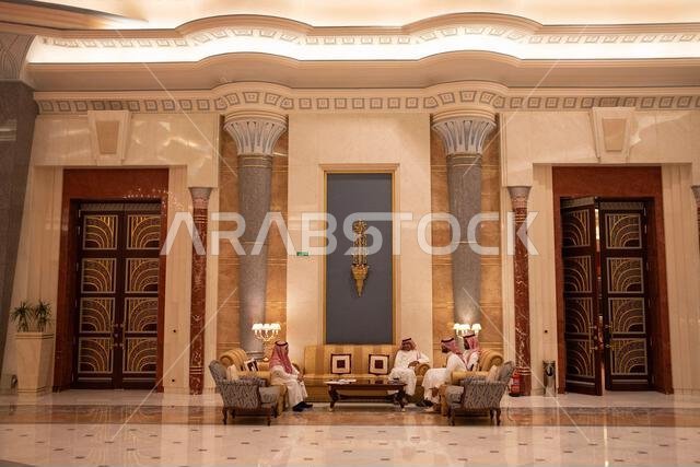 Three men in traditional attire sit on chairs in a grand lobby with marble floors and ornate columns.