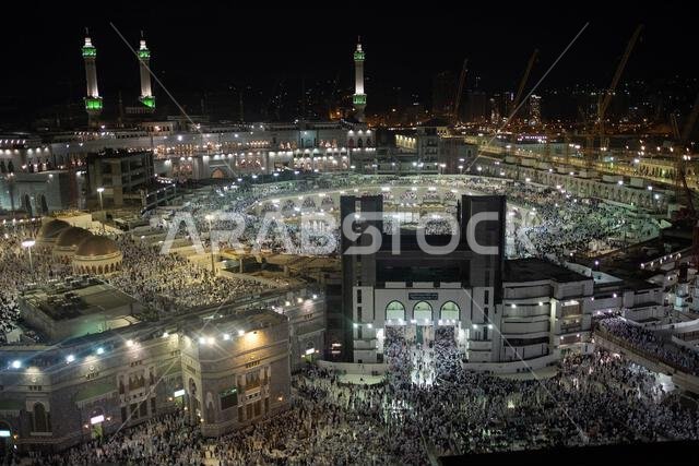 A large crowd of people gathers at night around a mosque with illuminated minarets and domes.