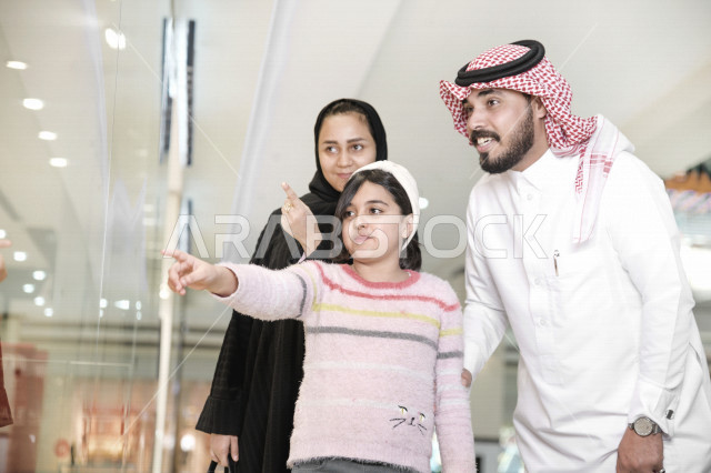 A young Saudi Arabian Gulf girl accompanied by her family pointing to something on the storefront with gestures indicating happiness, enjoying buying and shopping, having a good time shopping from the mall.