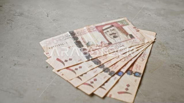أوراق نقدية من الريال السعودي منتشرة على سطح خرساني، تسلط الضوء على تفاصيل العملة المحلية في بيئة مالية بسيطة.