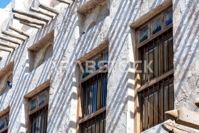 مبنى عربي تقليدي بنافذة خشبية ووسائد خشبية. يضفي الجدار المطلي باللون الأبيض والتفاصيل الخشبية الريفية إحساسًا بالتاريخ والثقافة.