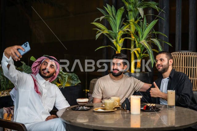 شباب عرب خليجيون سعوديون يجلسون في احد مقاهي المملكة العربية السعودية، شاب يحمل بيده الهاتف المحمول ويقوم بإلتقاط صورة سيلفي جماعية له و لأصدقائه، صورة جماعية تذكارية، تناول القهوة والمشروبات الباردة، أنشطة شبابية ترفيهية، الأجواء الشبابية في المقاهي السع