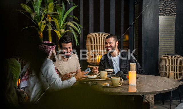 شباب عرب خليجيون سعوديون يجلسون في احد مقاهي المملكة العربية السعودية، شاب يحمل بيده الهاتف المحمول ويقوم بعرضه على أصدقائه للإستمتاع بمشاهدة البرامج الترفيهية، تناول القهوة والمشروبات الباردة، أنشطة شبابية ترفيهية، الأجواء الشبابية في المقاهي السعودية