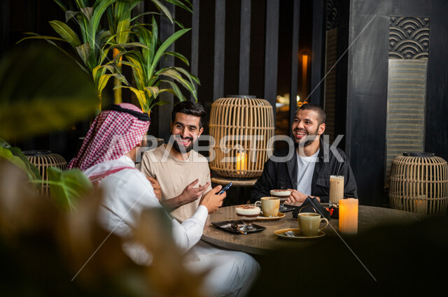 شباب عرب خليجيون سعوديون يجلسون في احد مقاهي المملكة العربية السعودية، شاب يحمل بيده الهاتف المحمول ويقوم بعرضه على أصدقائه للإستمتاع بمشاهدة البرامج الترفيهية، تناول القهوة والمشروبات الباردة، أنشطة شبابية ترفيهية، الأجواء الشبابية في المقاهي السعودية