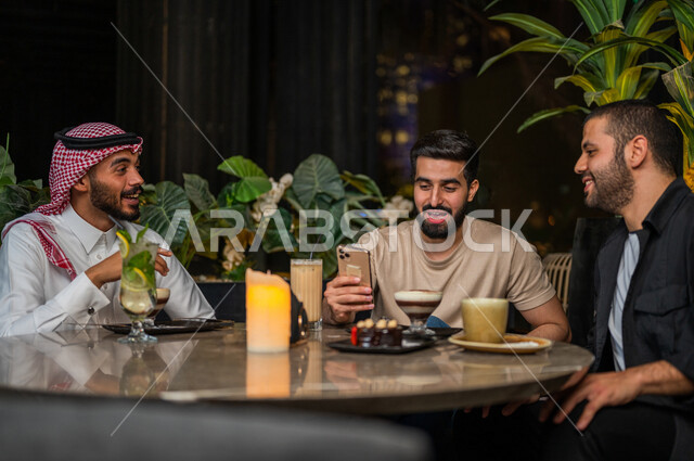 شباب عرب خليجيون سعوديون يجلسون في احد مقاهي المملكة العربية السعودية، شاب يحمل بيده الهاتف المحمول، الإستمتاع بمشاهدة البرامج الترفيهية، تناول القهوة والمشروبات الباردة، أنشطة شبابية ترفيهية، الأجواء الشبابية في المقاهي السعودية