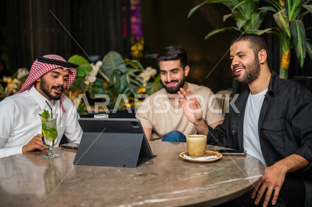 شباب عرب خليجيون سعوديون يجلسون في احد مقاهي المملكة العربية السعودية، اجراءة مكالمة فيديو من خلال التابليت المحمول، القاء التحية من خلال شاشة التابليت، بث فيديو مباشر، دردشة وتواصل، ايماءات وجه ويدين تدل على السعادة، الأجواء الشبابية في المقاهي السعودية