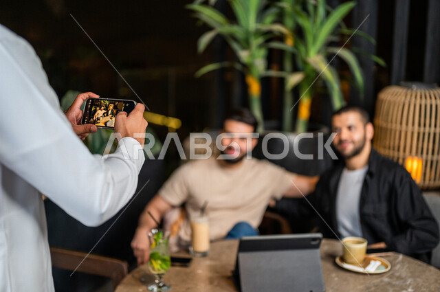 شباب عرب خليجيون سعوديون يجلسون في احد مقاهي المملكة العربية السعودية وامامهم جهاز التابليت المحمول، شاب يحمل بيده الهاتف المحمول يقوم بإلتقاط صورة جماعية له ولأصدقائه بسعادة، صورة جماعية تذكارية، أنشطة شبابية ترفيهية، الأجواء الشبابية في المقاهي السعودية