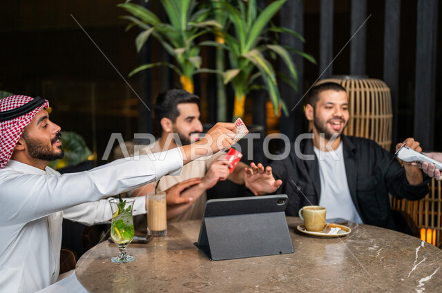 شباب عرب خليجيون سعوديون يجلسون في احد مقاهي المملكة العربية السعودية وامامهم جهاز التابليت، شاب يحاول دفع حساب المشروبات بدل من اصدقائه، استخدام البطاقة الائتمانية في دفع الحساب، المشروبات الباردة، الأجواء الشبابية في المقاهي السعودية