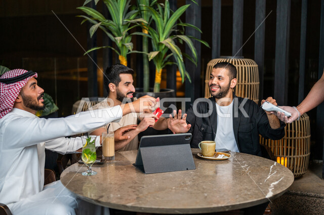 شباب عرب خليجيون سعوديون يجلسون في احد مقاهي المملكة العربية السعودية وامامهم جهاز التابليت، شاب يحاول دفع حساب المشروبات بدل من اصدقائه، استخدام البطاقة الائتمانية في دفع الحساب، المشروبات الباردة، الأجواء الشبابية في المقاهي السعودية