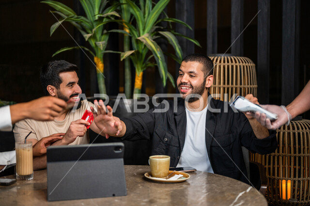 شباب عرب خليجيون سعوديون يجلسون في احد مقاهي المملكة العربية السعودية وامامهم جهاز التابليت، شاب يحاول دفع حساب المشروبات بدل من اصدقائه، استخدام البطاقة الائتمانية في دفع الحساب، المشروبات الباردة، الأجواء الشبابية في المقاهي السعودية