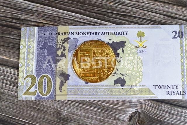 عملة الريال السعودي الورقية مزودة بعملة بيتكوين ذهبية مادية، بيتكوين هي أول عملة مشفرة لامركزية، وعقد في شبكة بيتكوين من نظير إلى نظير للتحقق من المعاملات