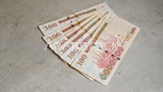 أوراق نقدية سعودية من فئة 100 ريال موزعة بشكل مروحي على سطح خرساني، مما يبرز العملة الشرق أوسطية في إطار مالي واضح.