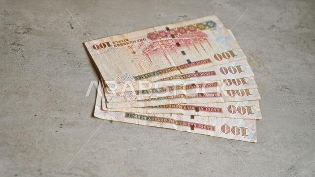 أوراق نقدية سعودية من فئة 100 ريال موضوعة على سطح خرساني، لعرض العملة والموضوع الاقتصادي في بيئة بسيطة.