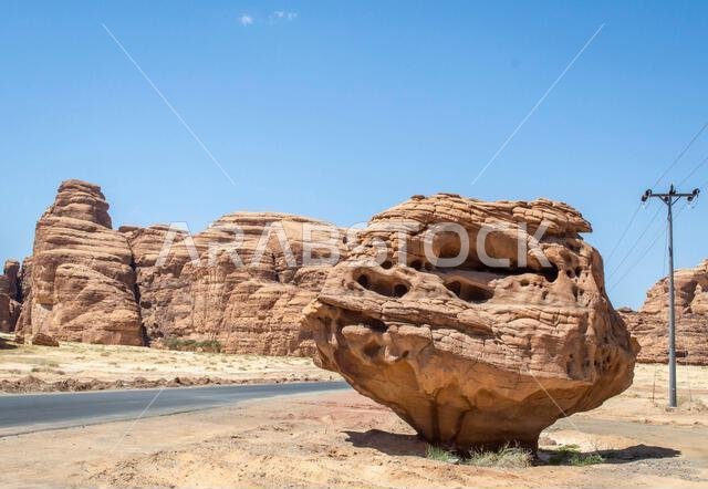 منظر لصخرة كبيرة على جانب الطريق في مدينة العلا السعودية، مع الجبال الصخرية في الخلفية في يوم مشمس.