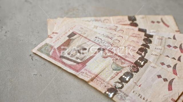 أوراق نقدية من الريال السعودي مبعثرة على سطح خرساني رمادي، تُظهر التصميم التفصيلي والعملة السعودية.