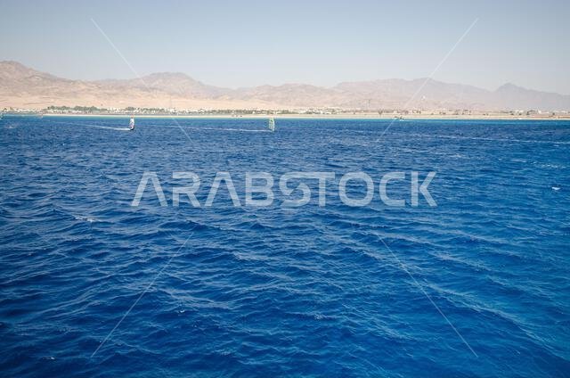 منظر بانورامي للبحر الأحمر بأمواجه الزرقاء المتلألئة تحت سماء صافية. تصطف الجبال البعيدة على الأفق، لتخلق تباينًا لافتًا. مشهد بحري هادئ.